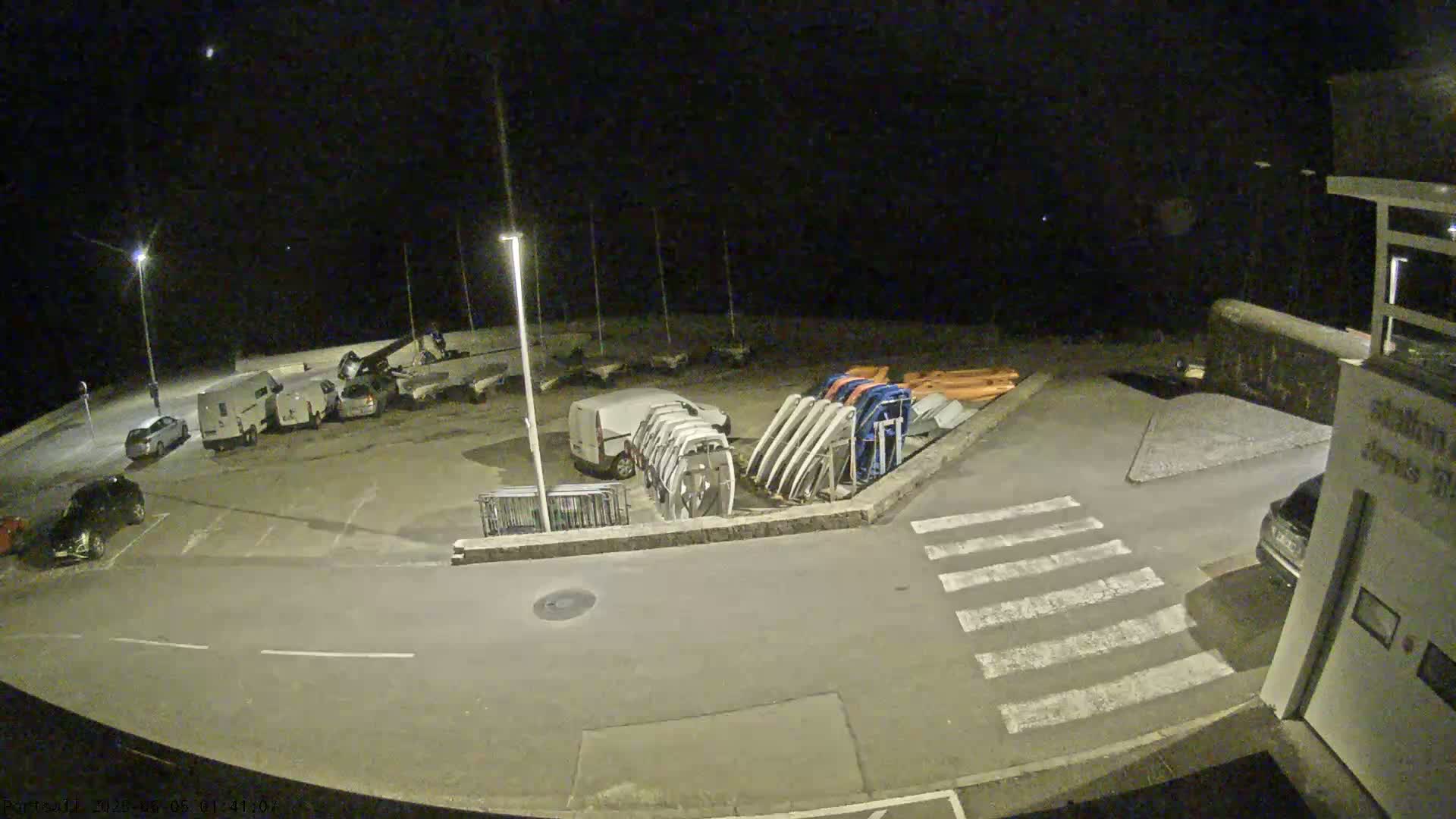 A nighttime, outdoor view of a parking lot with several cars and vans, boat trailers, and other equipment near a crosswalk.