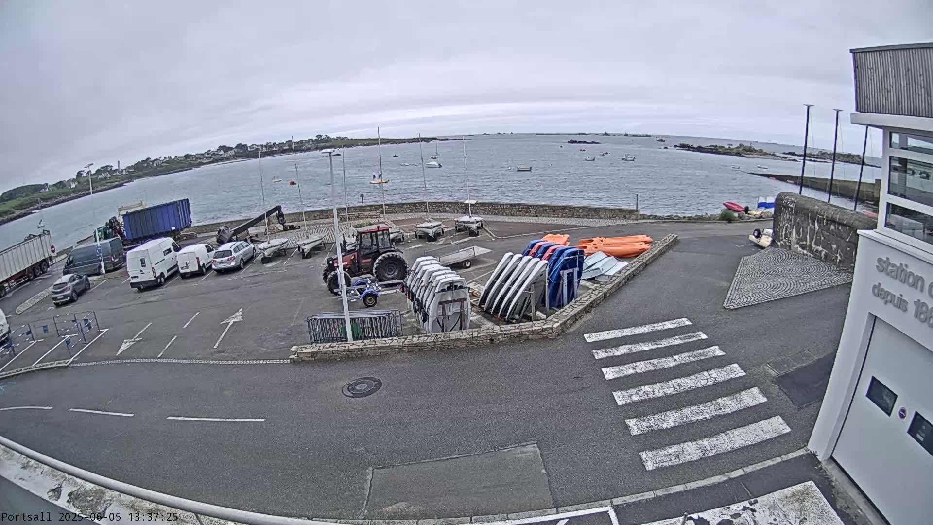 A parking lot with several vehicles, a tractor, and stacks of small boats is situated next to a calm body of water under an overcast sky.