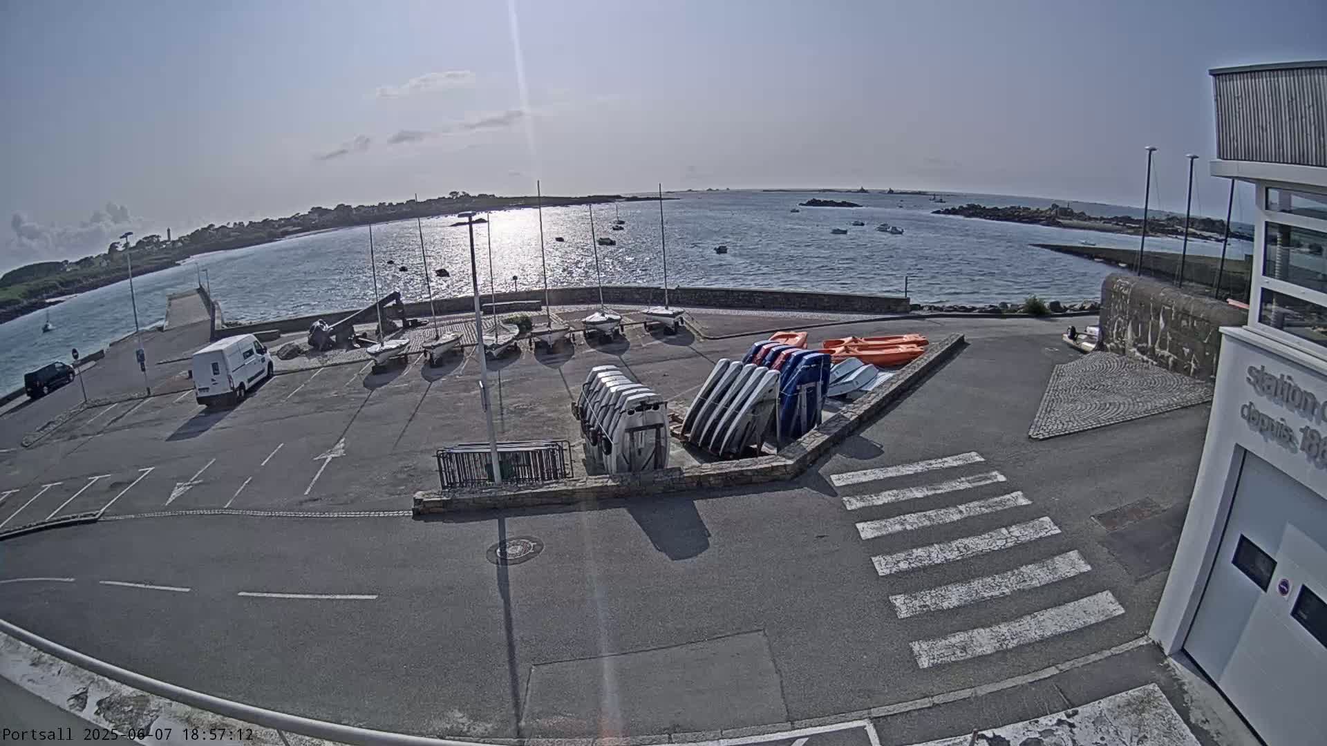 A sunny day at a harbor shows a parking lot with several small sailboats and kayaks stored near a building, overlooking calm water with distant land.