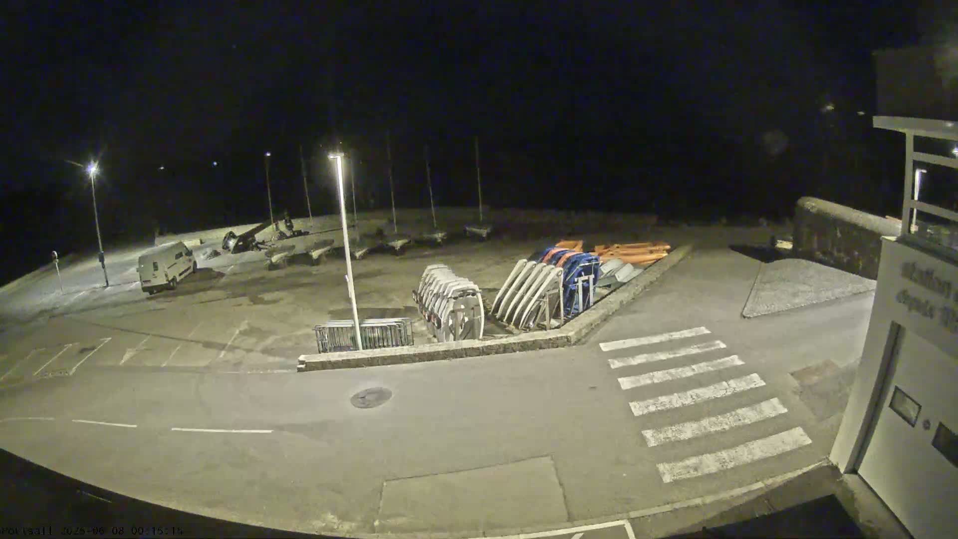 A nighttime, outdoor view shows a mostly empty parking lot with several small boats and a van, next to a building and a crosswalk.