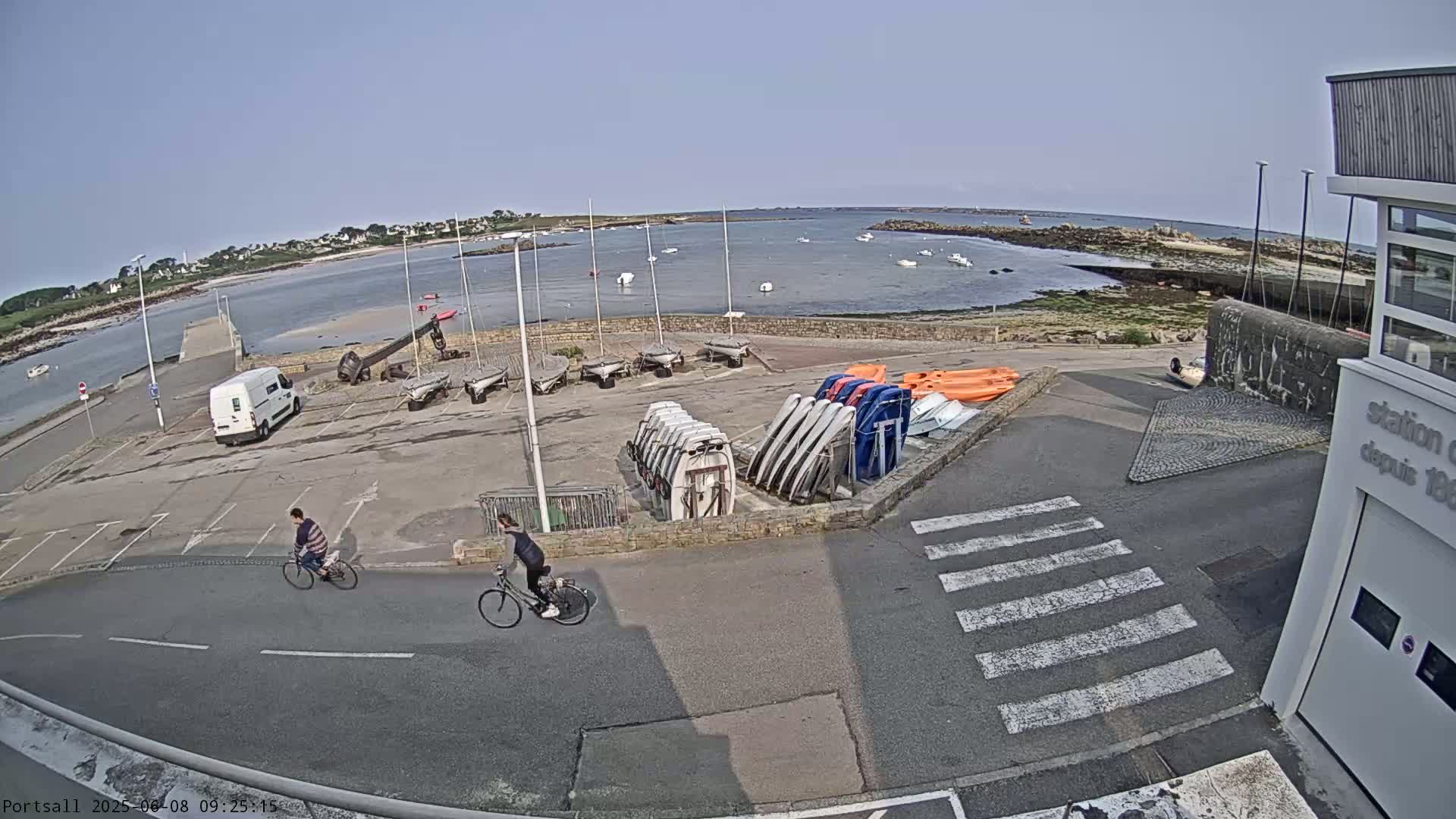 A sunny day at a harbor shows numerous small sailboats and other watercraft stored on land, with two people riding bicycles on a nearby road.