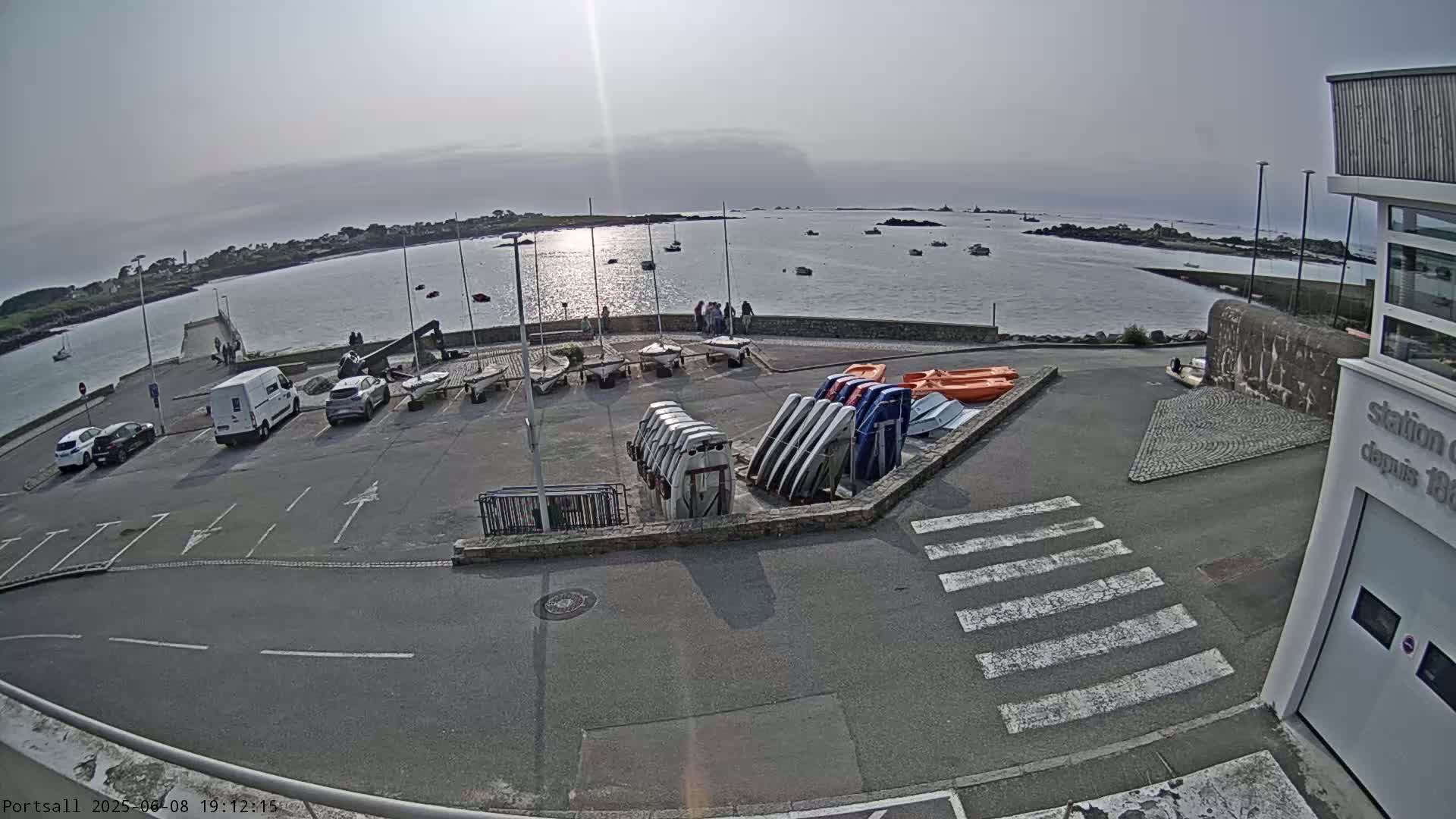 A parking lot with several cars and numerous sailboats is situated near a body of water under a mostly sunny sky.