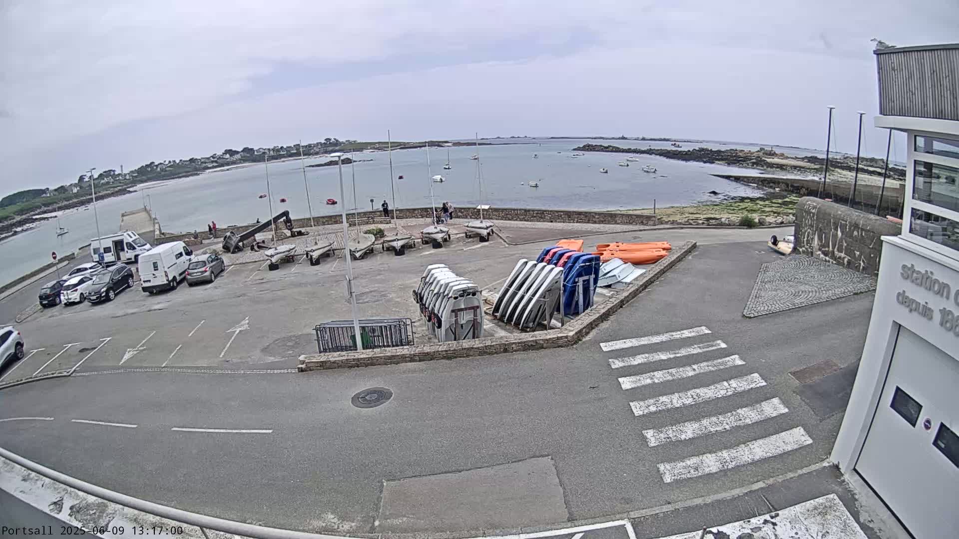 A parking lot with several cars and numerous sailboats and kayaks stored on racks overlooks a calm body of water under an overcast sky.