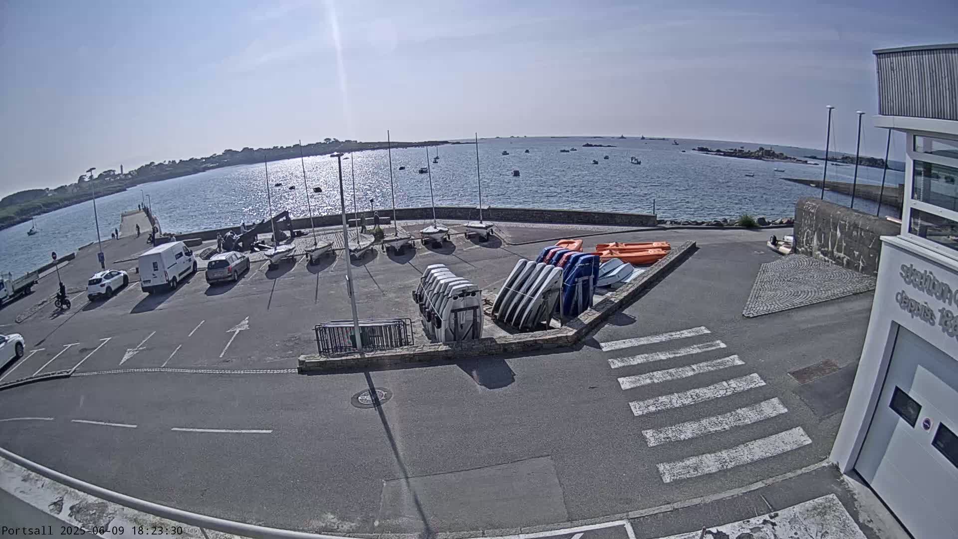A sunny day at a harbor shows several parked cars, sailboats, and small motorboats near a calm body of water with islands in the distance.