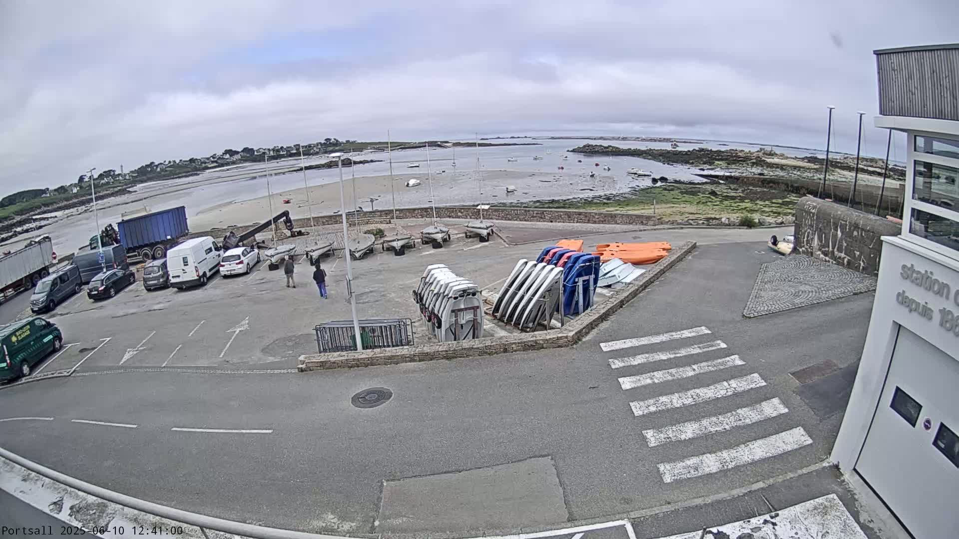 A parking lot with several cars and trucks is next to a boat storage area overlooking a calm bay under an overcast sky.