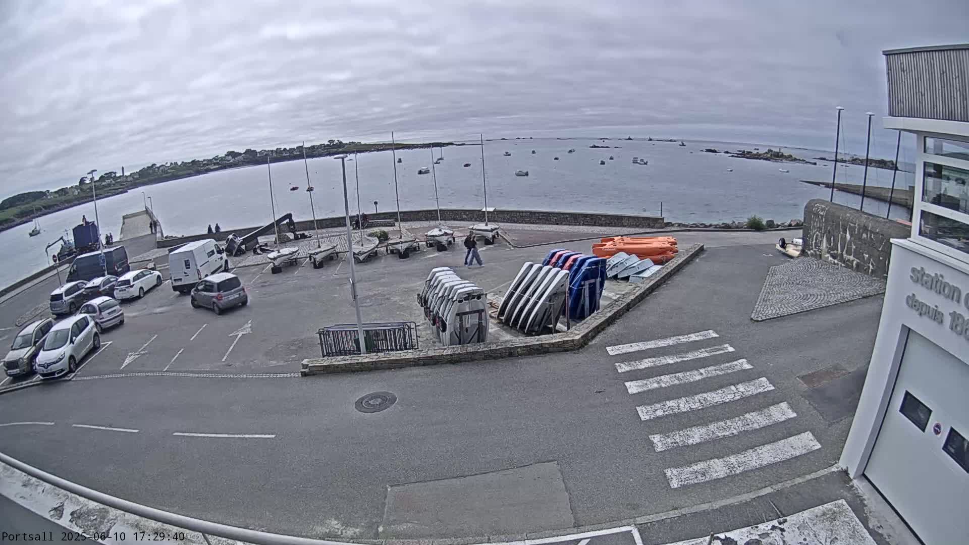 A parking lot with several cars and numerous small boats and sailboats is situated next to a calm body of water under an overcast sky.