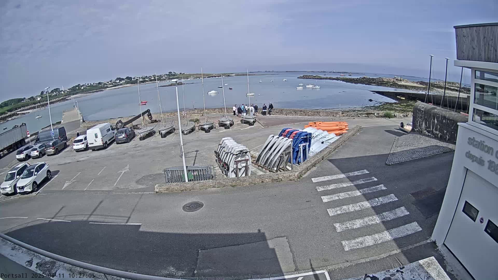 A sunny day at a harbor shows a parking lot filled with cars and trailers, next to a collection of small sailboats and kayaks, all overlooking a calm body of water with several small boats and islands in the distance.