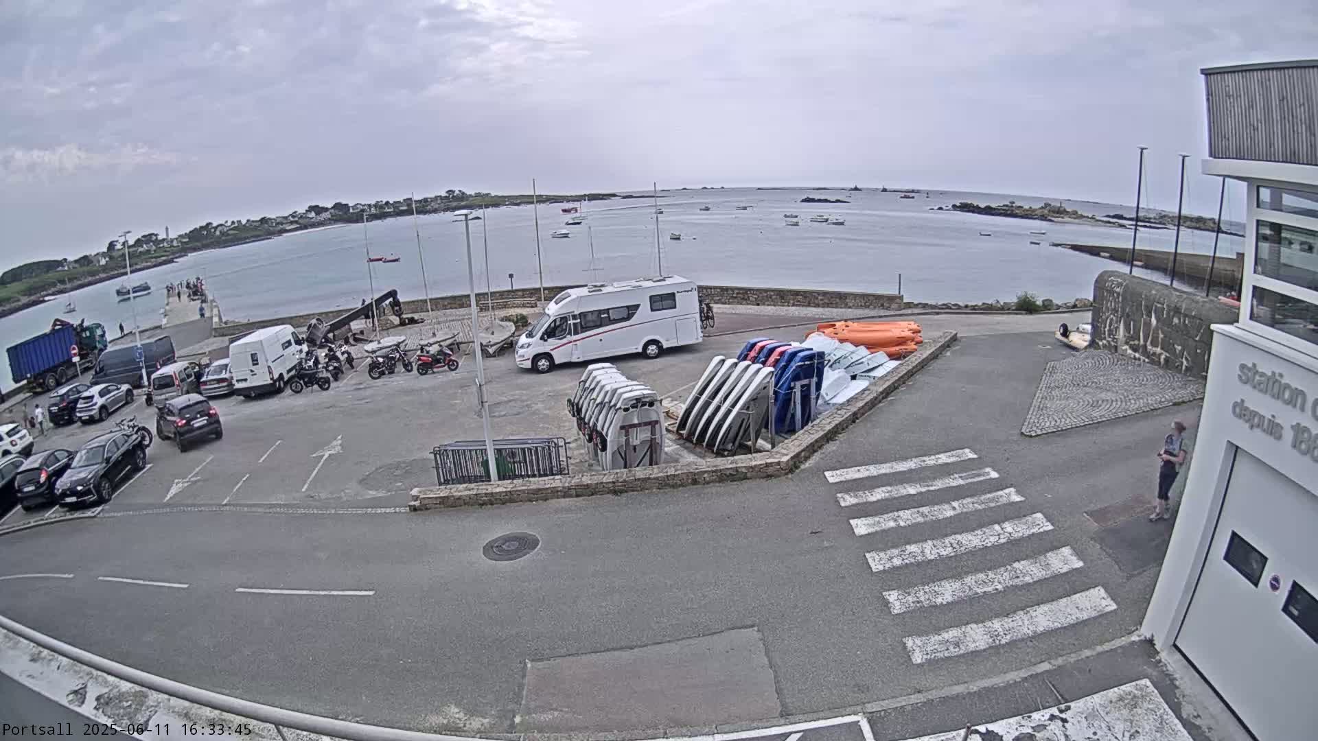 A parking lot with cars, motorcycles, a campervan, and several boats is situated beside a calm body of water under an overcast sky.