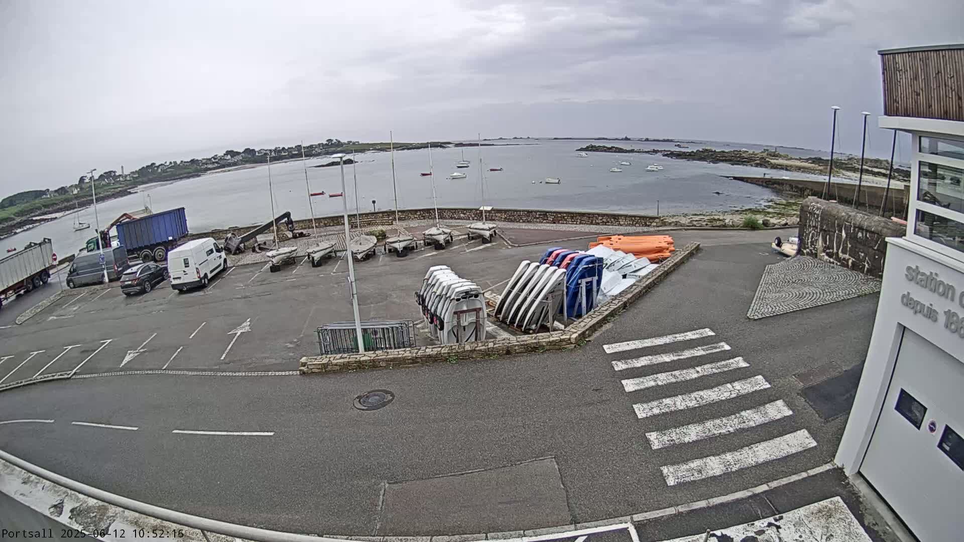 A parking lot with several vehicles and numerous small sailboats and dinghies near a calm body of water under an overcast sky.