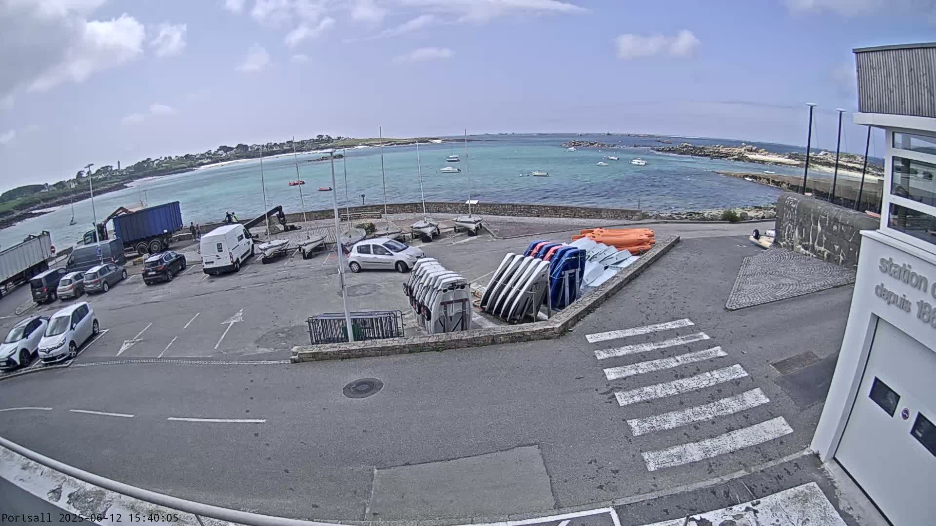 A parking lot with cars and boats is next to a calm body of water under a partly cloudy sky.