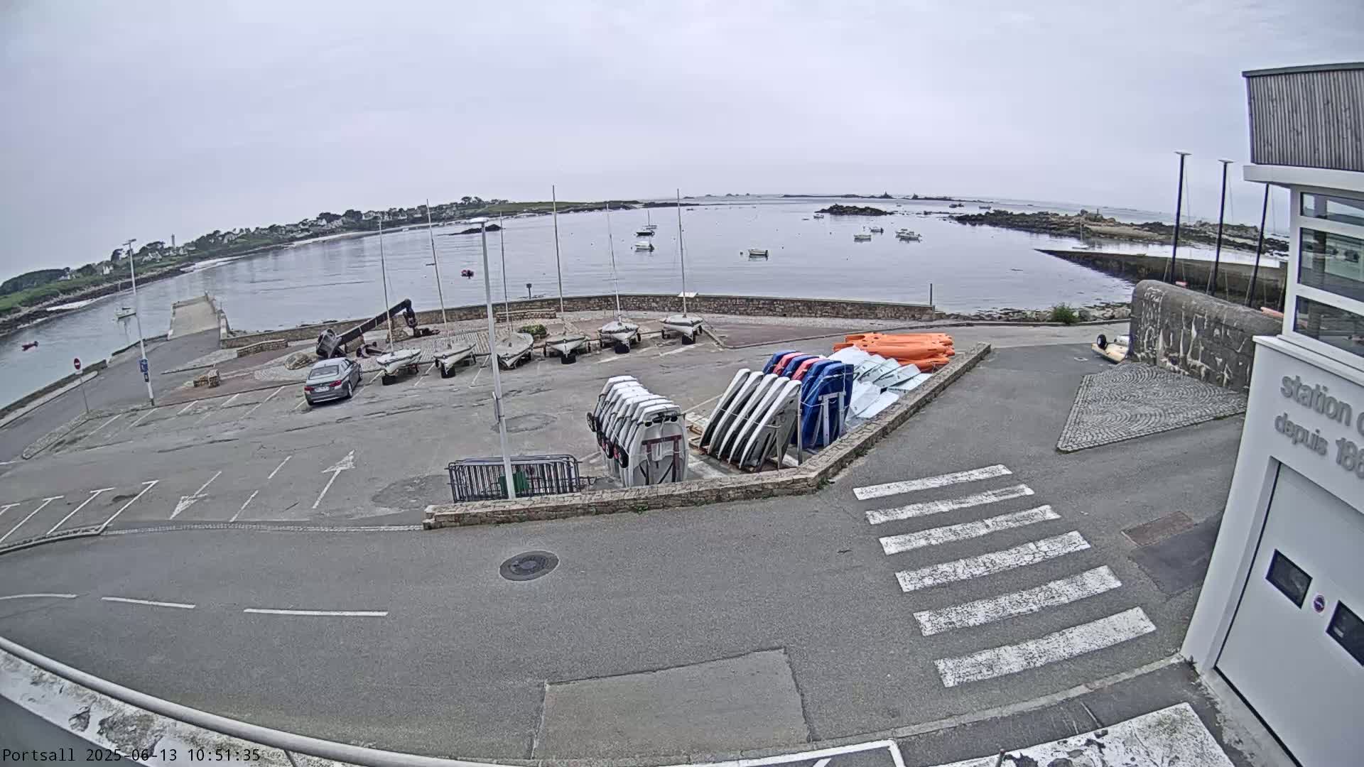 A parking lot with several sailboats and small boats stored on racks overlooks a calm body of water under an overcast sky.