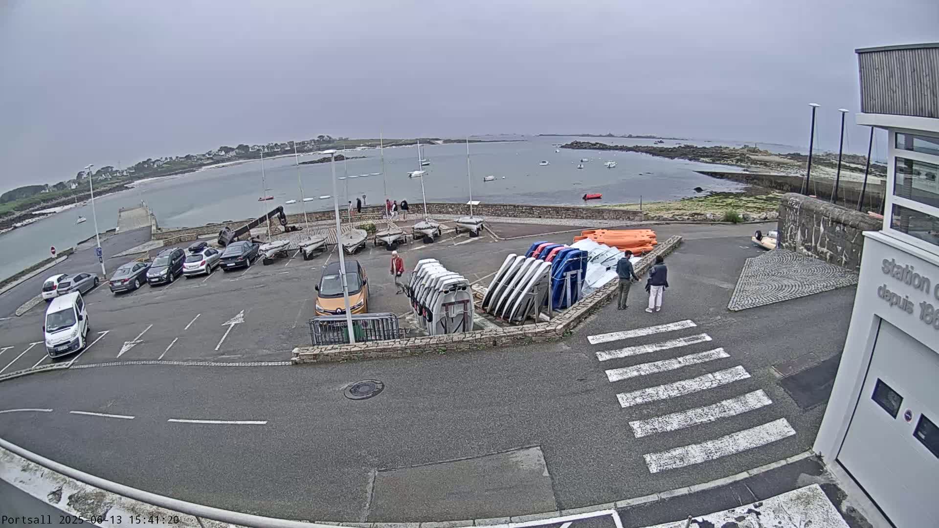 A parking lot with numerous cars and several small boats on racks overlooks a calm body of water on an overcast day.