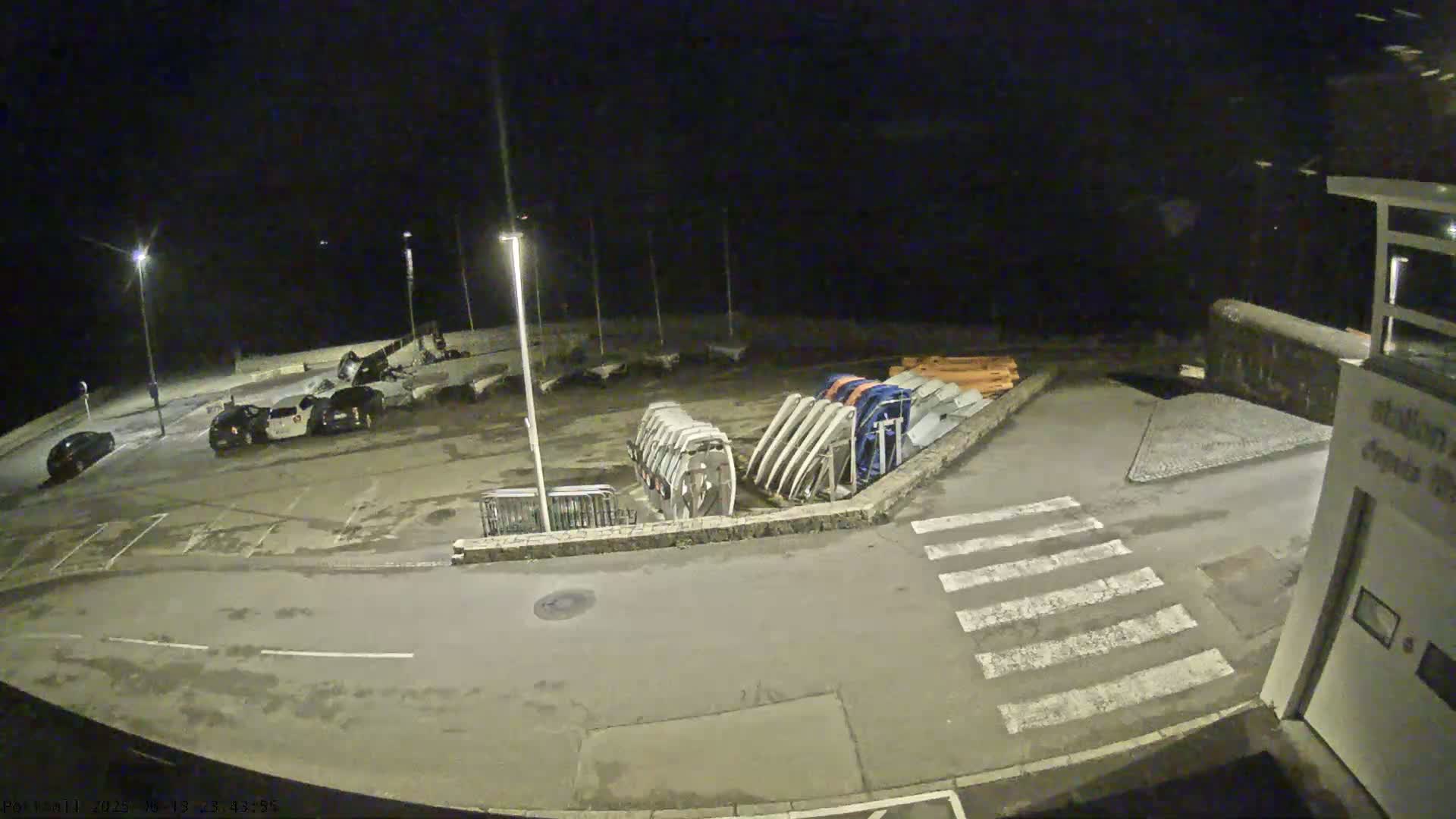 A nighttime, outdoor view of a mostly empty parking lot next to a harbor with several boats and boat parts visible under artificial light.