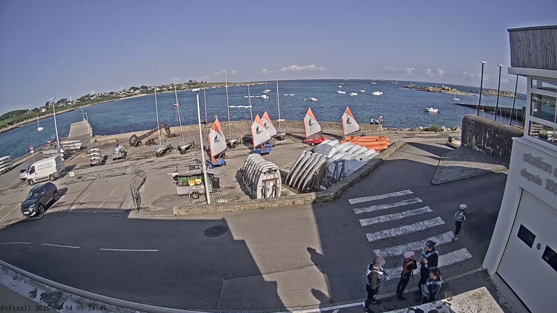 A harbor scene on a sunny day shows sailboats and small boats in the water and numerous small sailboats and dinghies stored on land near a building and a parking area with several vehicles.