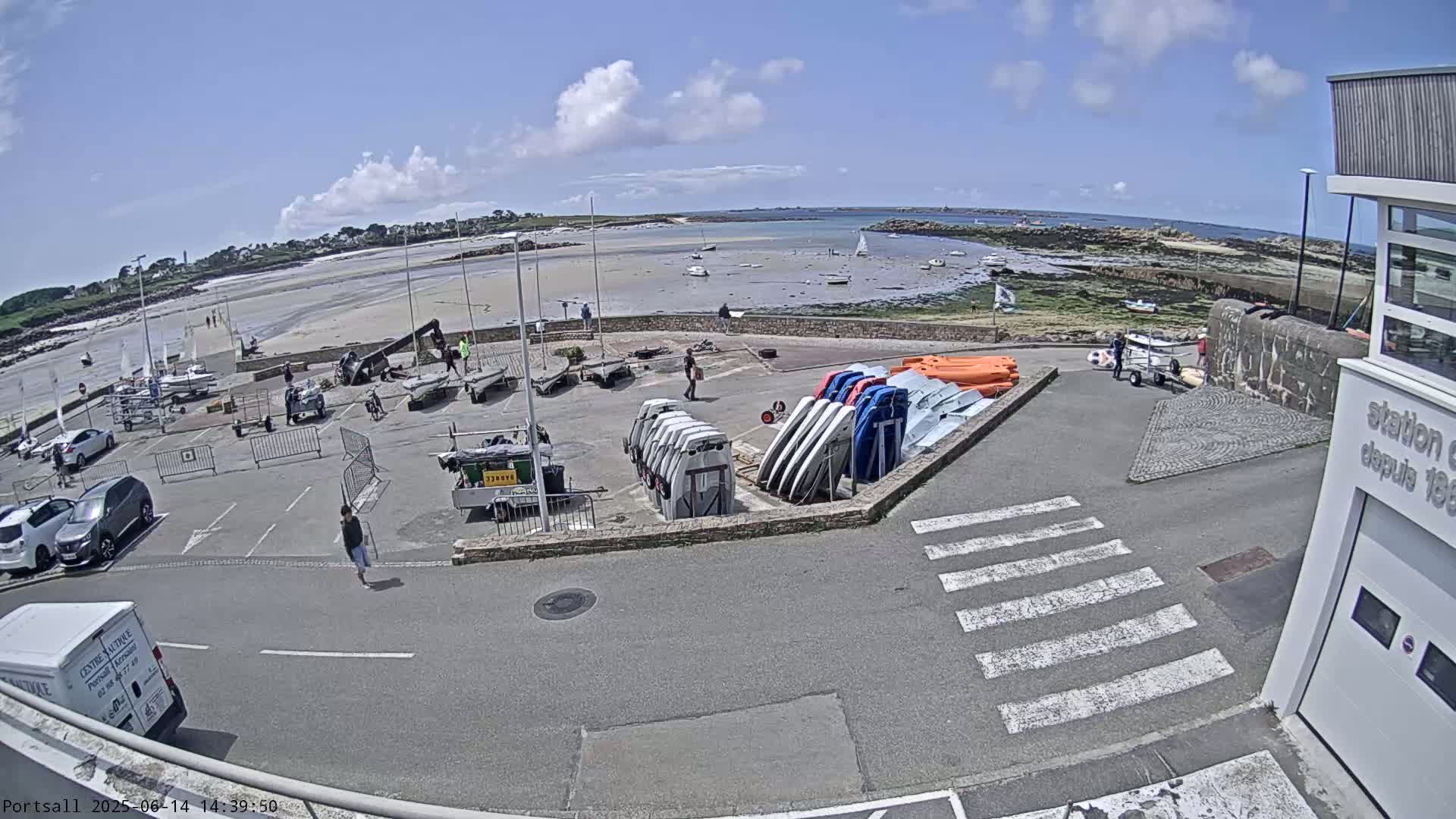 A sunny day at a harbor shows several sailboats on a sandy beach, with numerous small boats and vehicles parked in a lot near a building.