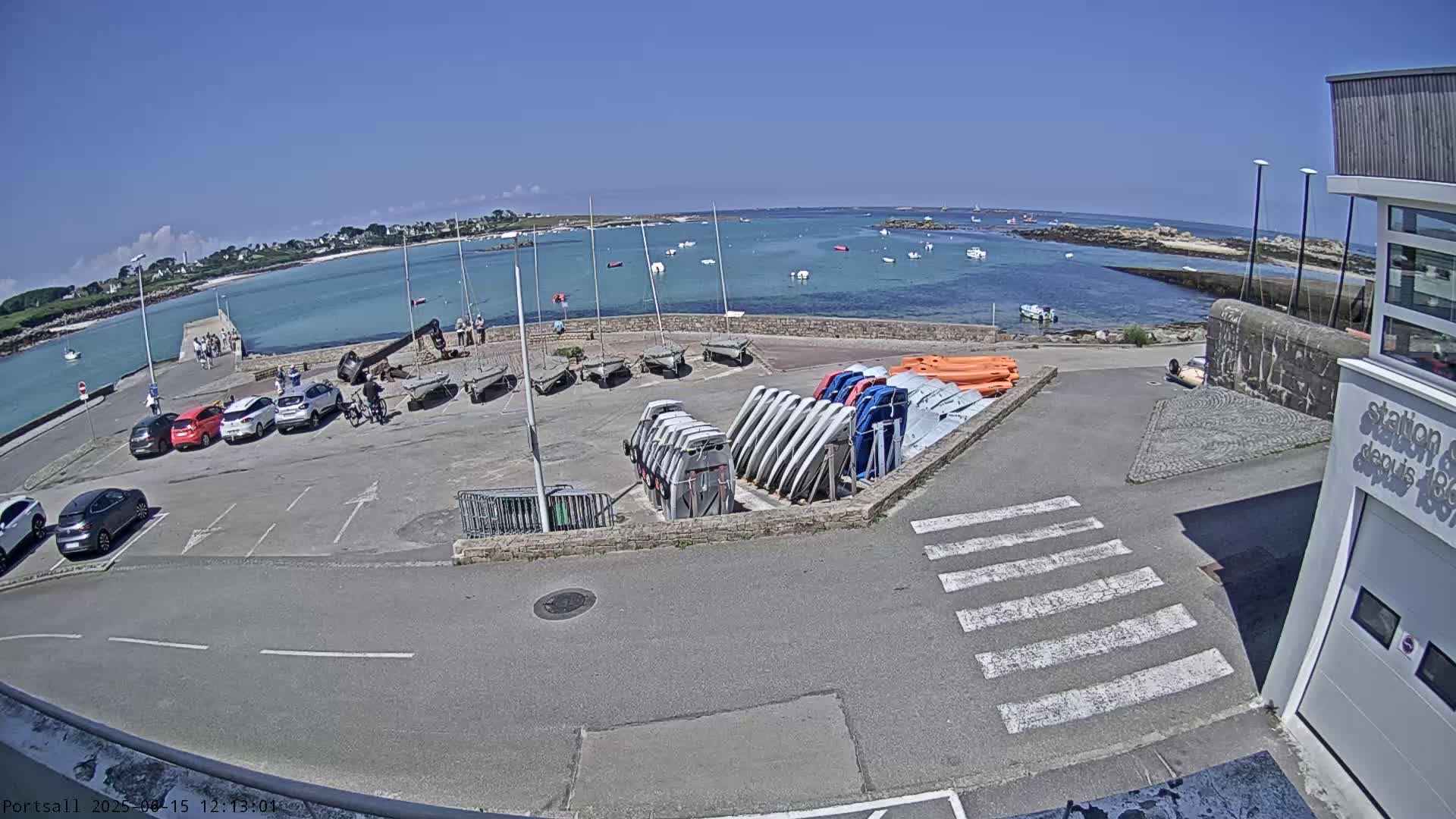 A sunny day at a harbor shows parked cars, several small sailboats stored upright, and a calm body of water with more boats anchored in the distance.