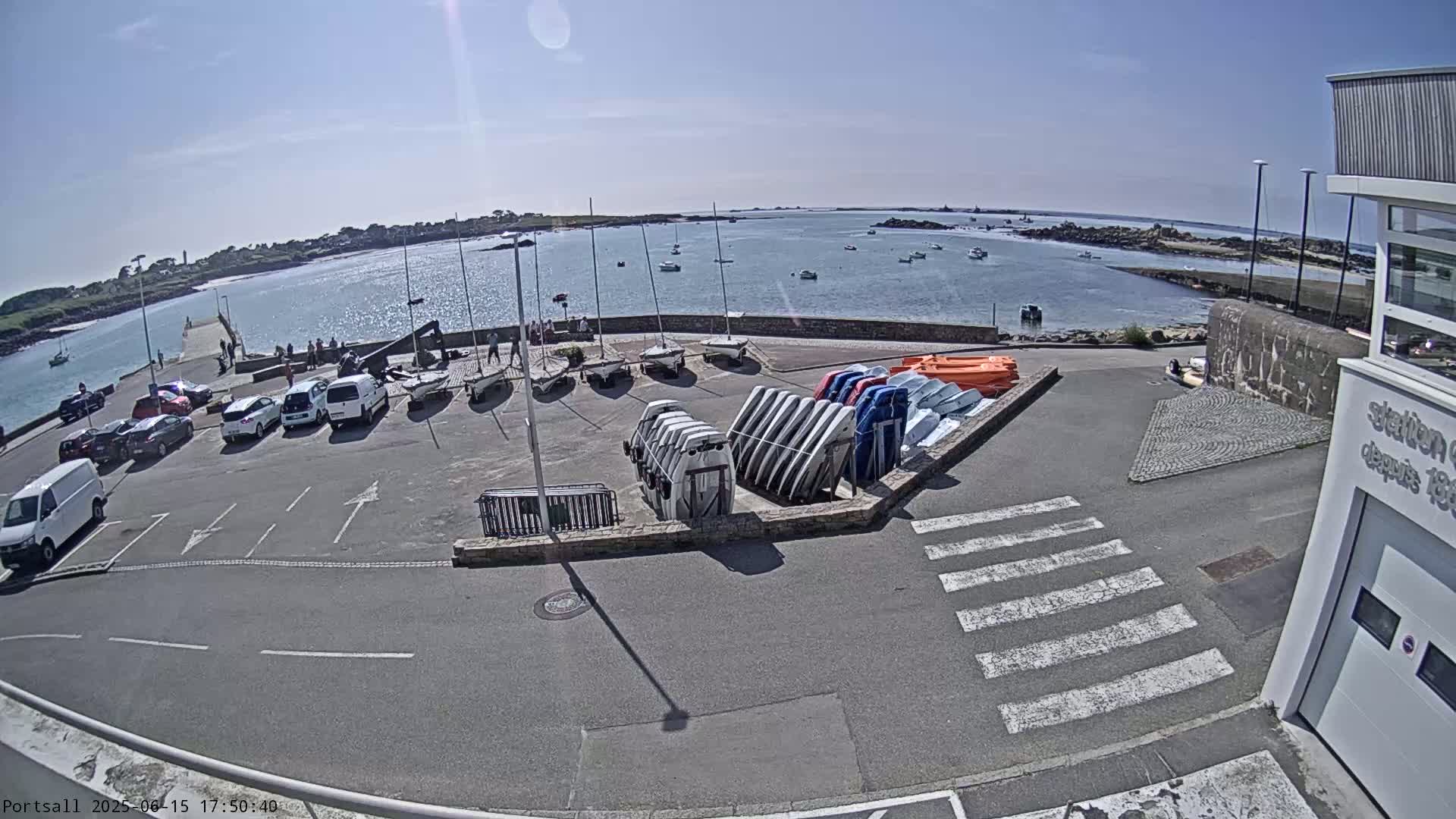 A sunny day at a harbor shows sailboats docked, many more stored upright, and cars parked in a lot next to a building.