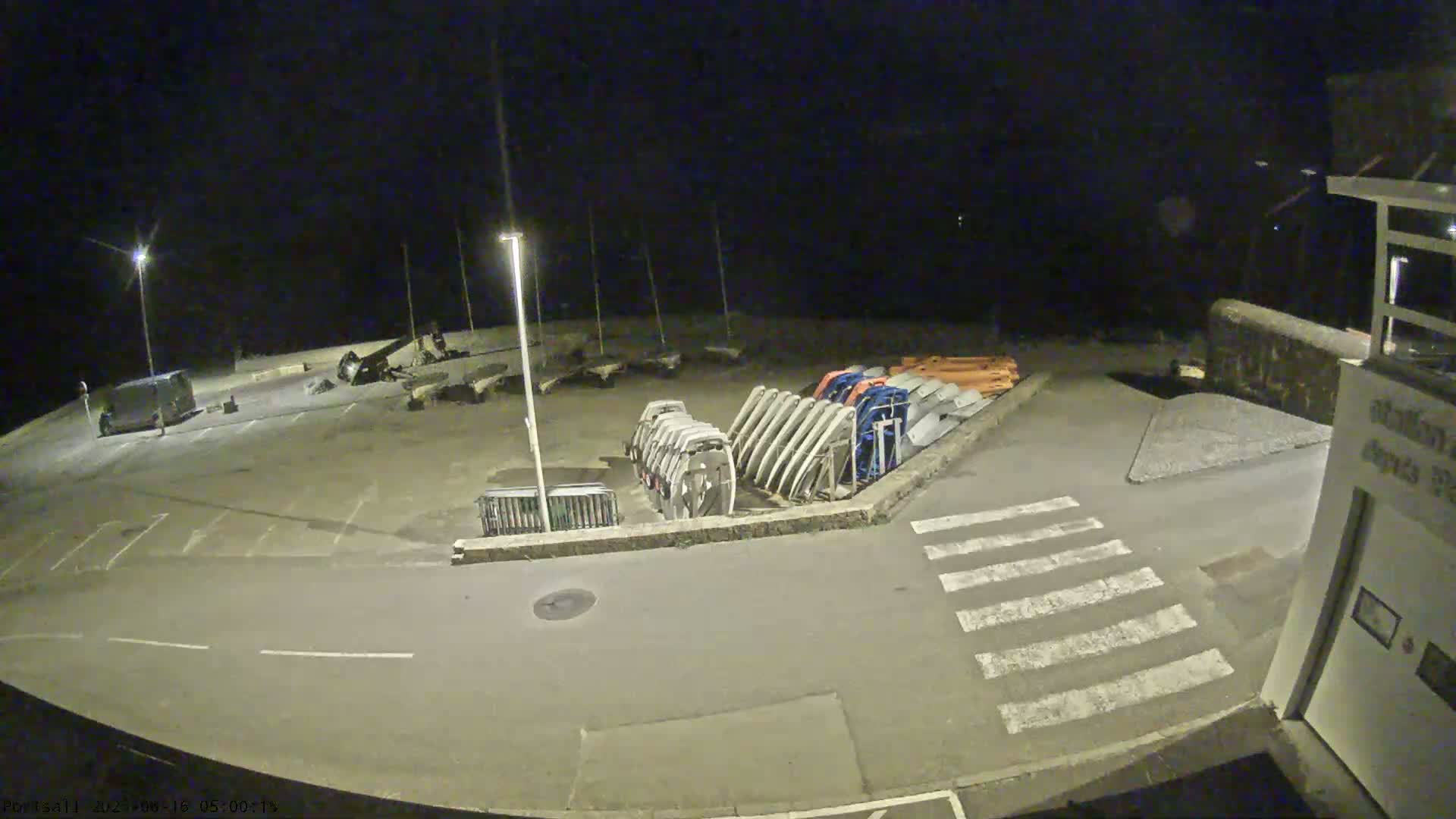A nighttime, empty parking lot next to a harbor with several boats and dinghies stored upside down.