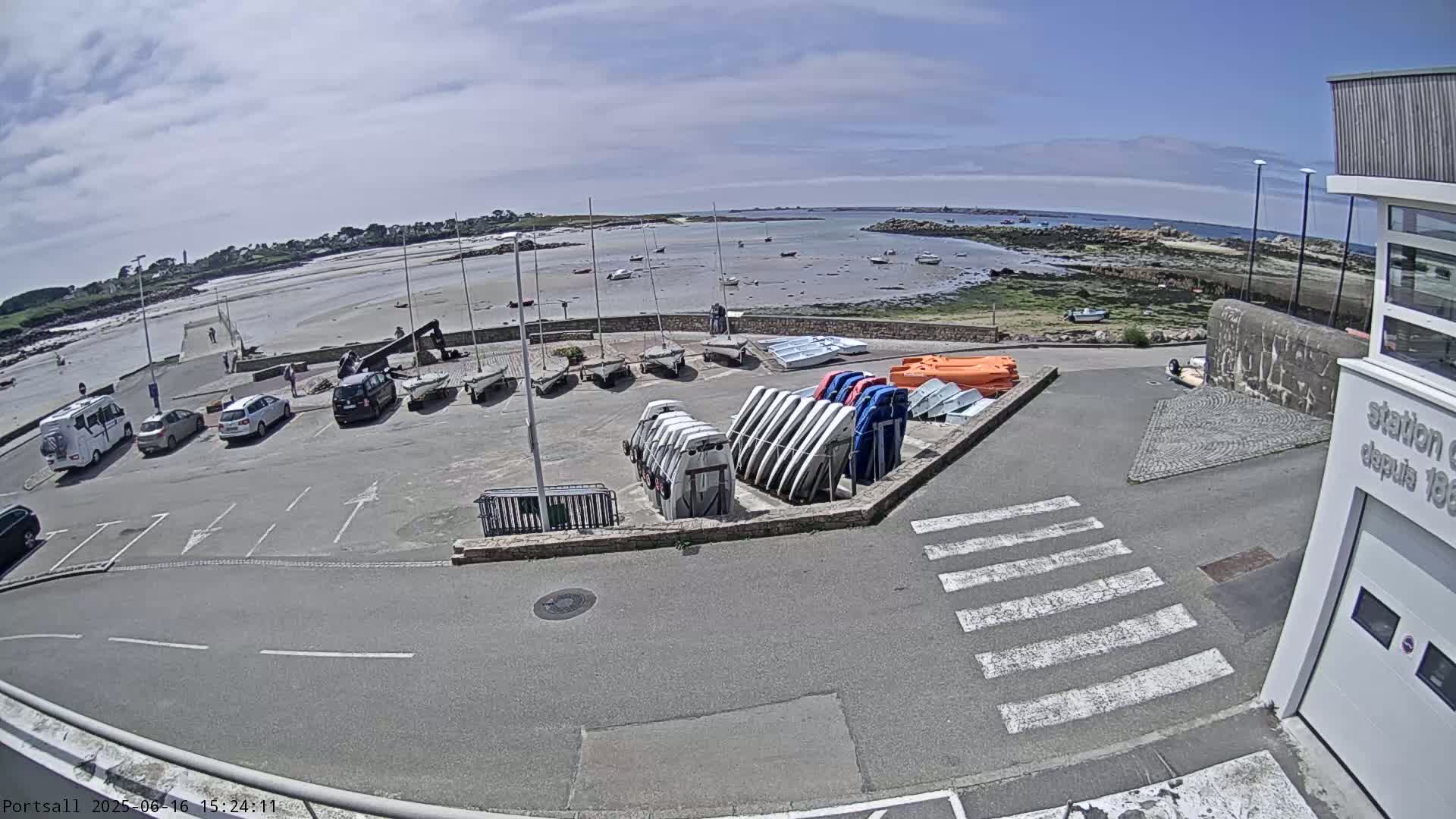 A parking lot with several cars and numerous small boats stored on racks overlooks a sandy beach and calm water under a partly cloudy sky.