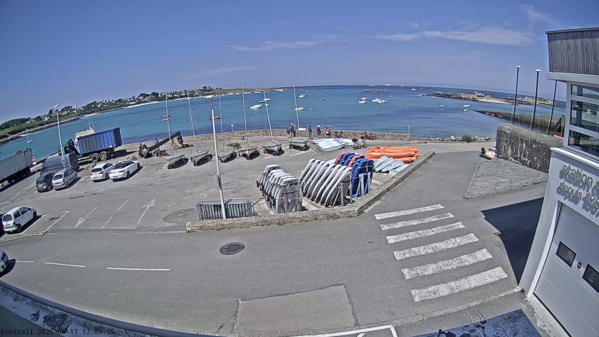 A sunny day overlooking a harbor with parked cars, boats, and sailboats,  a building, and a few people.