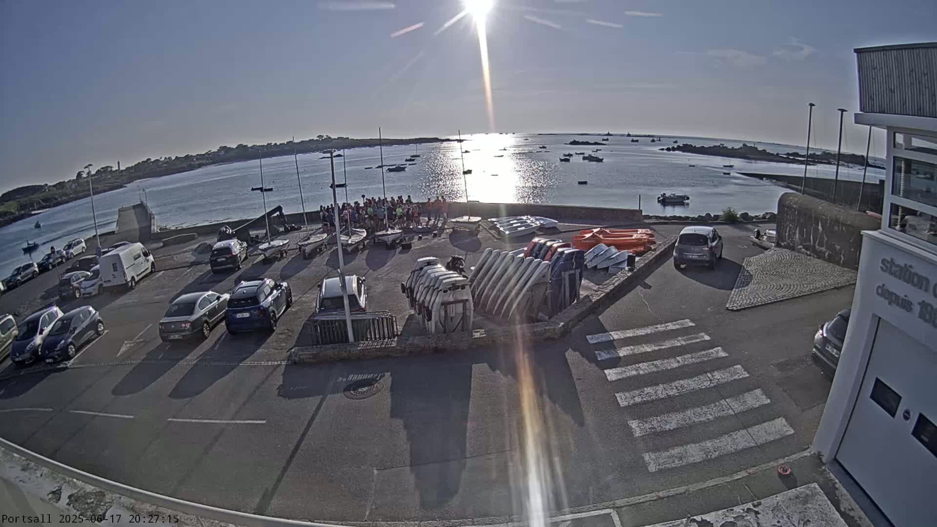A sunny day at a harbor shows a parking lot full of cars near a collection of sailboats and other watercraft, with a group of people standing near the water's edge.