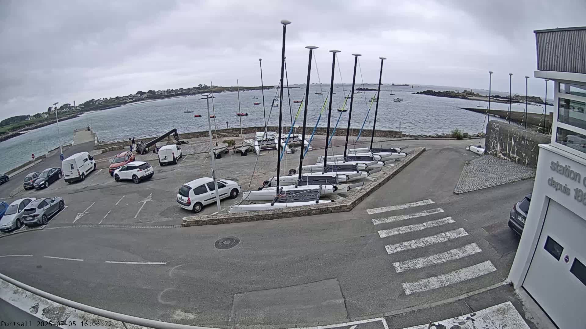 A parking lot with several cars and a row of sailboats is situated beside a calm body of water under an overcast sky.