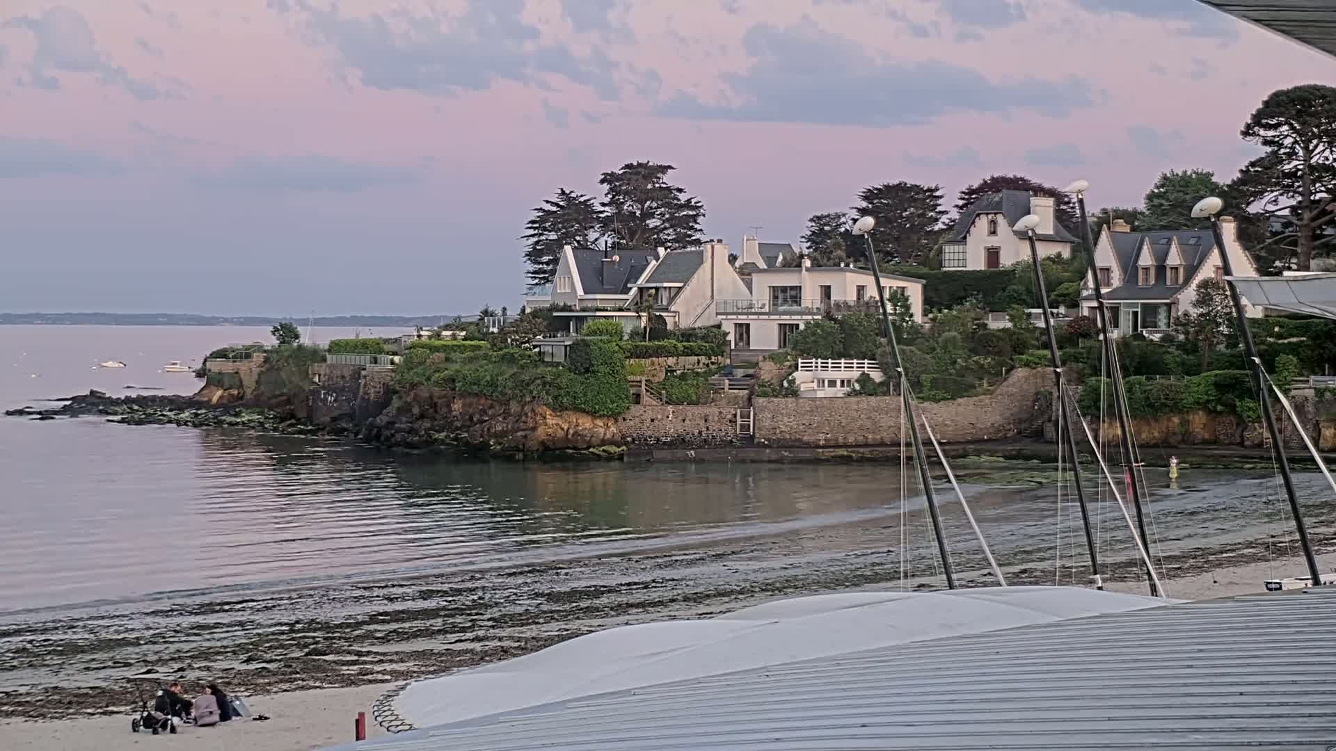 A tranquil coastal scene at sunset shows several white houses perched atop a rocky cliff overlooking a calm bay with a sandy beach and several sailboats moored in shallow water.