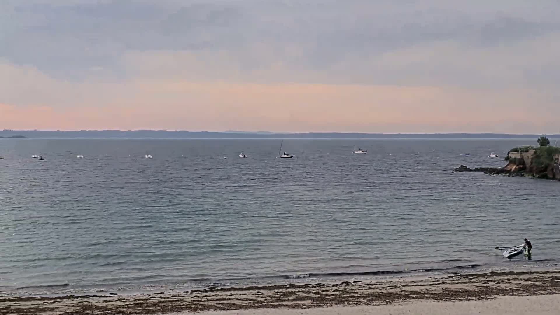 A calm, overcast day shows a sandy beach with a person preparing a small boat near a rocky shoreline, with several small boats visible in the distance on the water.