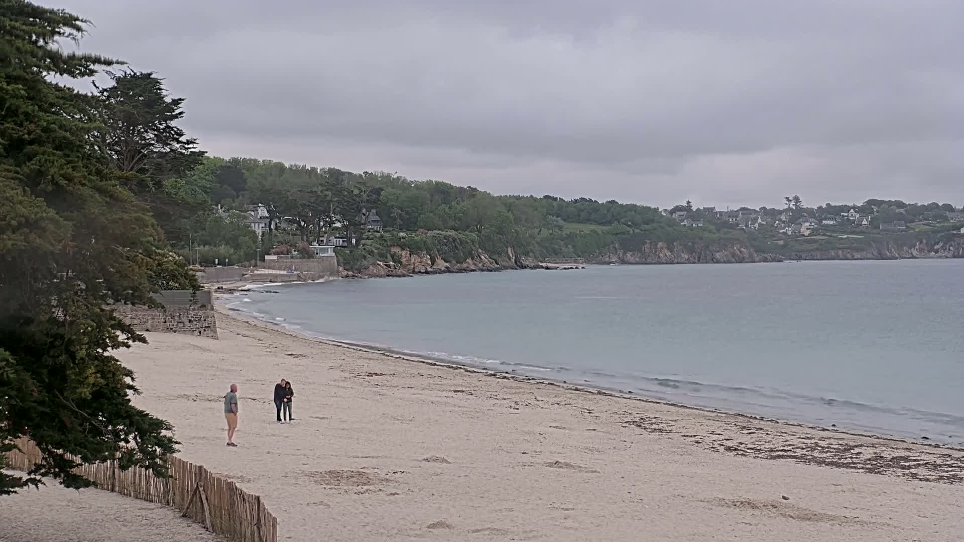 A sandy beach with a few people on it stretches along a calm, light-blue ocean under an overcast sky, backed by green cliffs and houses.