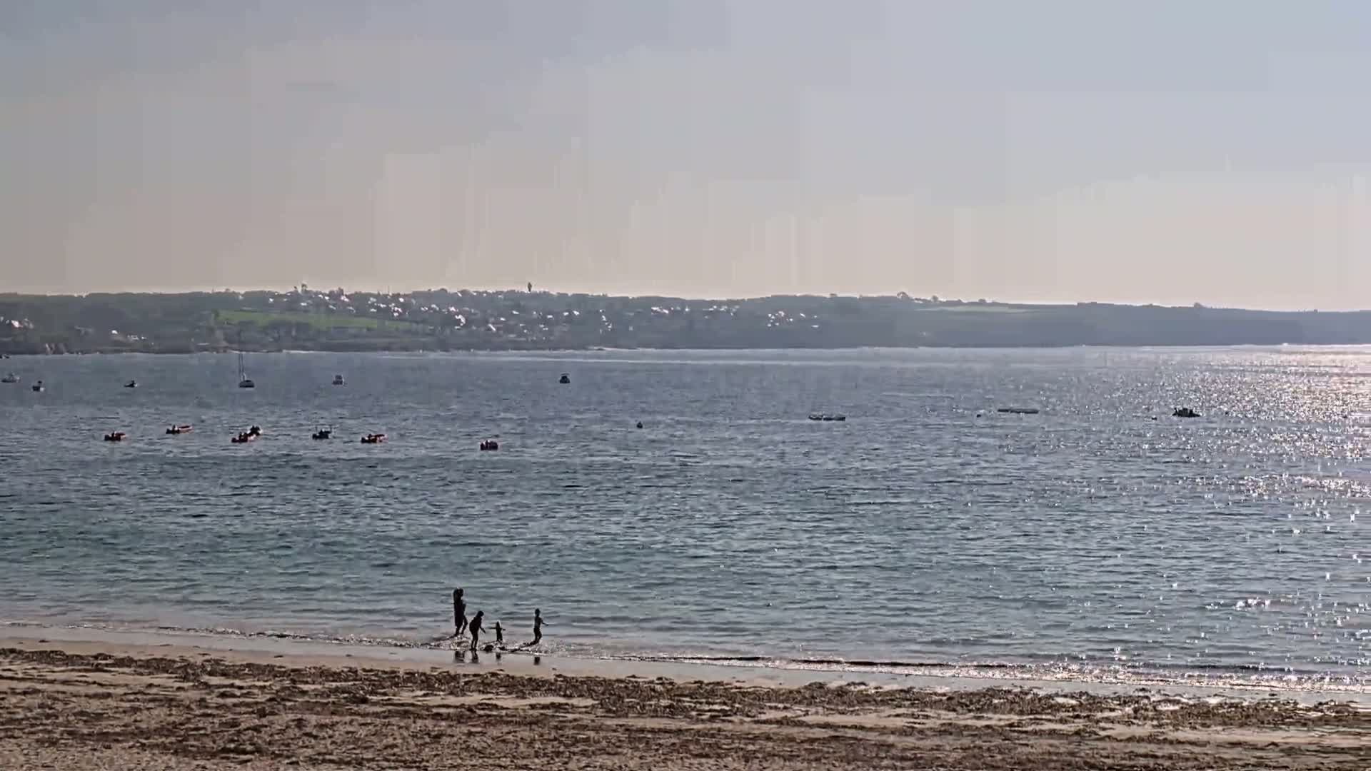 A sandy beach with a family playing in the shallows of a calm, sunlit sea, with several boats visible in the distance.