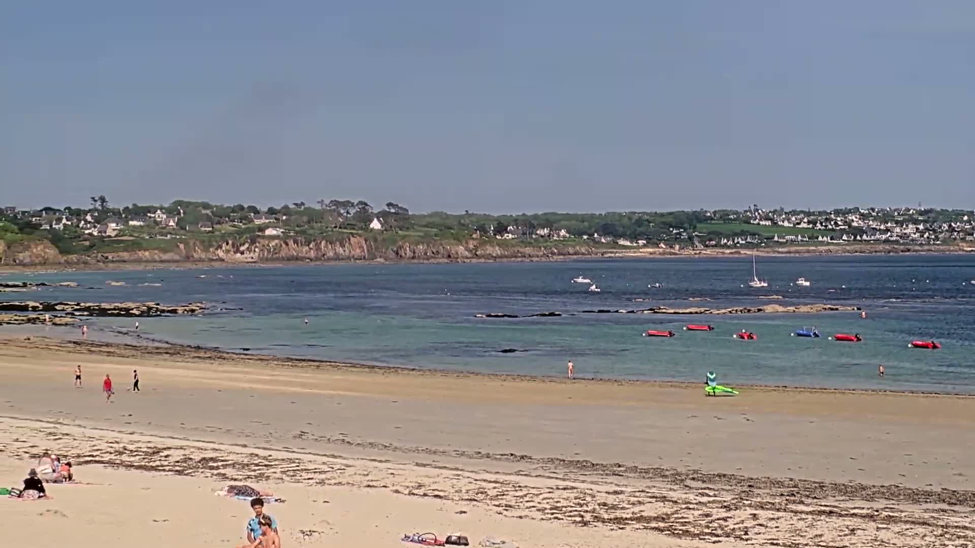 A sandy beach with people sunbathing and swimming in calm, clear water, under a sunny sky, overlooks a bay with several small boats and a distant town.