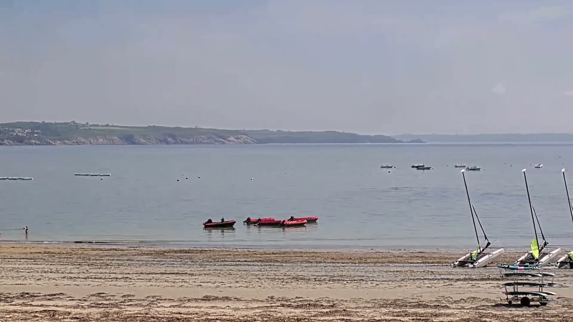 A sandy beach with several small boats and sailboats on the shore and in calm, clear water, with a distant coastline visible under a hazy sky.