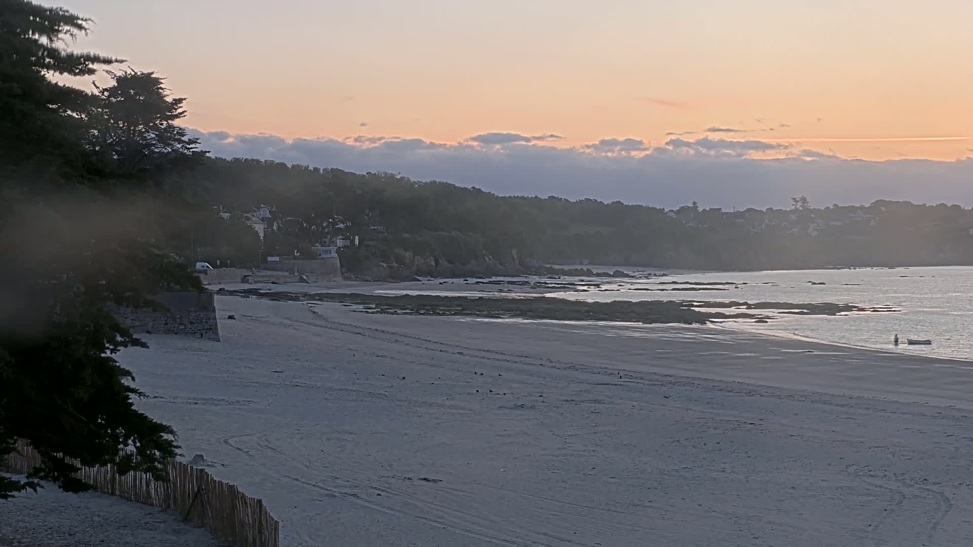 A sandy beach stretches to a calm ocean at sunset, with a rocky shoreline and tree-lined hills in the background.