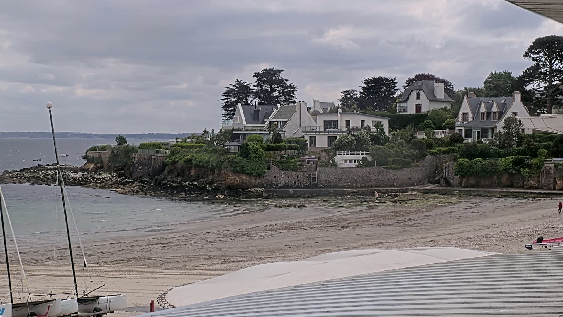 A sandy beach with two sailboats rests near a rocky shoreline where several white houses sit atop a lush, green hill under an overcast sky.