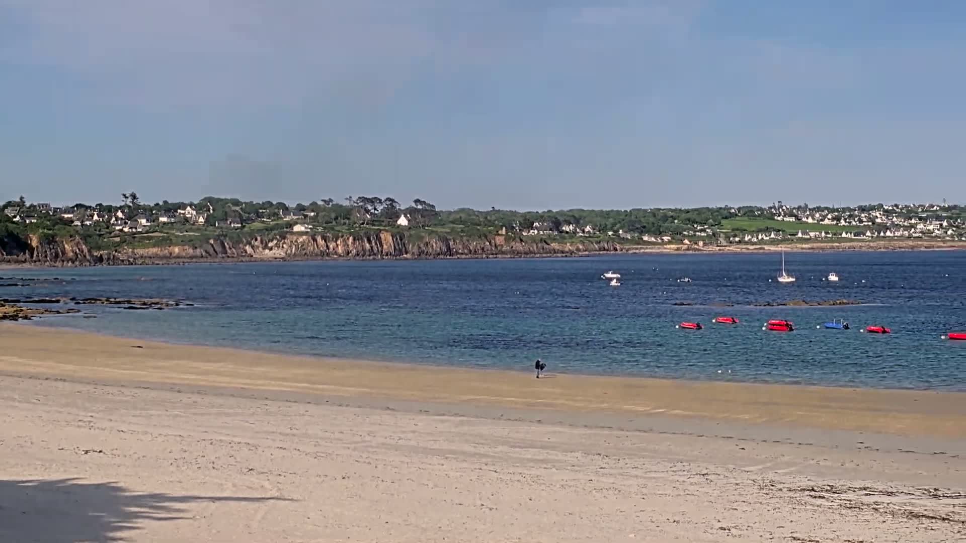A sandy beach stretches toward a calm bay with several small boats and a distant coastline under a clear sky.