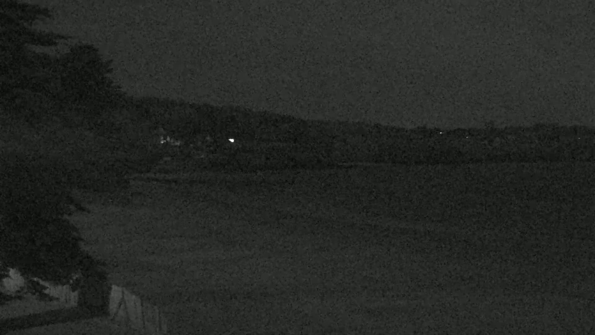 A dark, nighttime scene shows a body of water in the distance, with a few faint lights visible on the opposite shore and some dark trees to the left.
