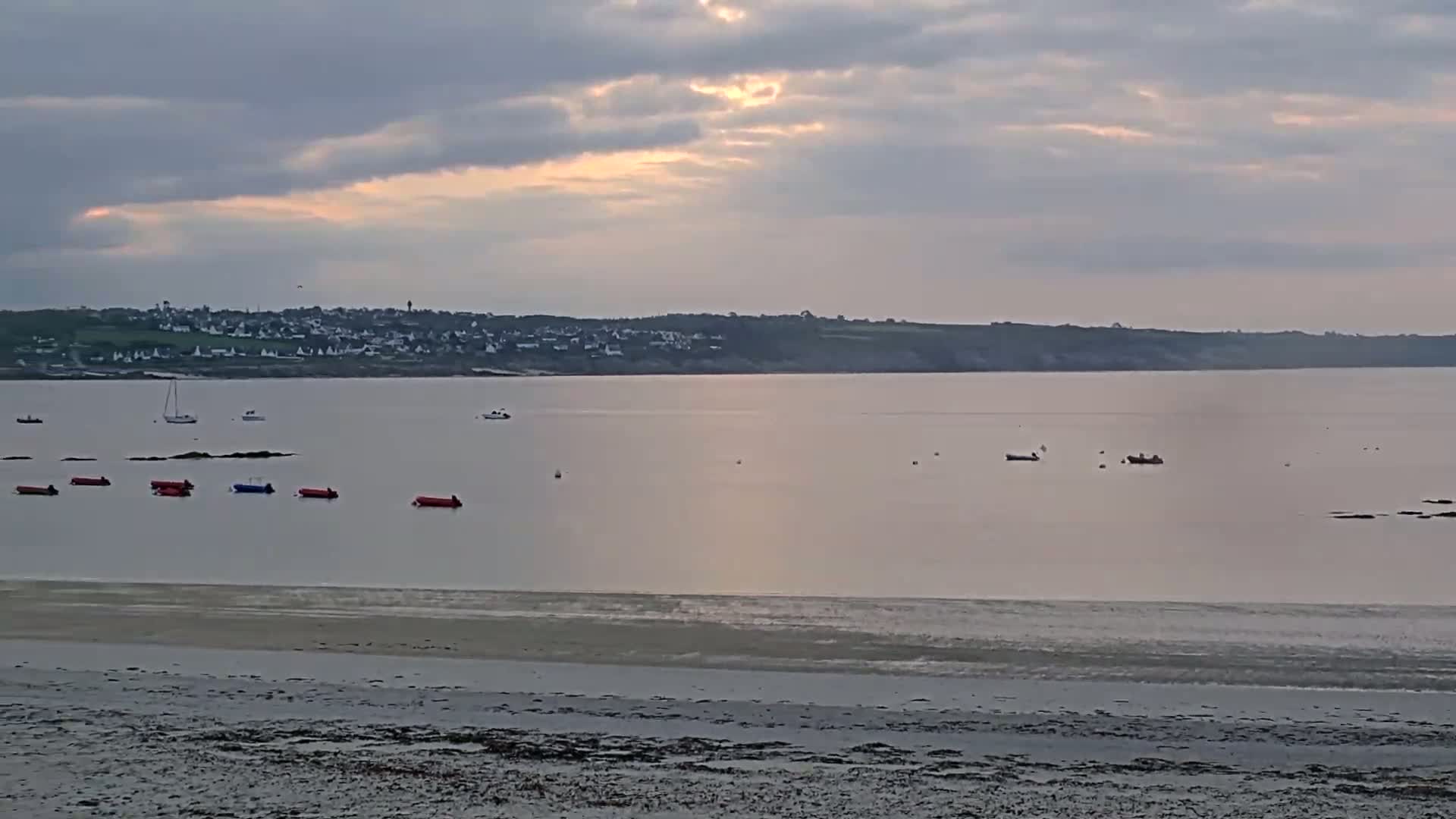 A sandy beach stretches towards a calm body of water where several small boats are scattered, under a cloudy sky with a hint of sunset.