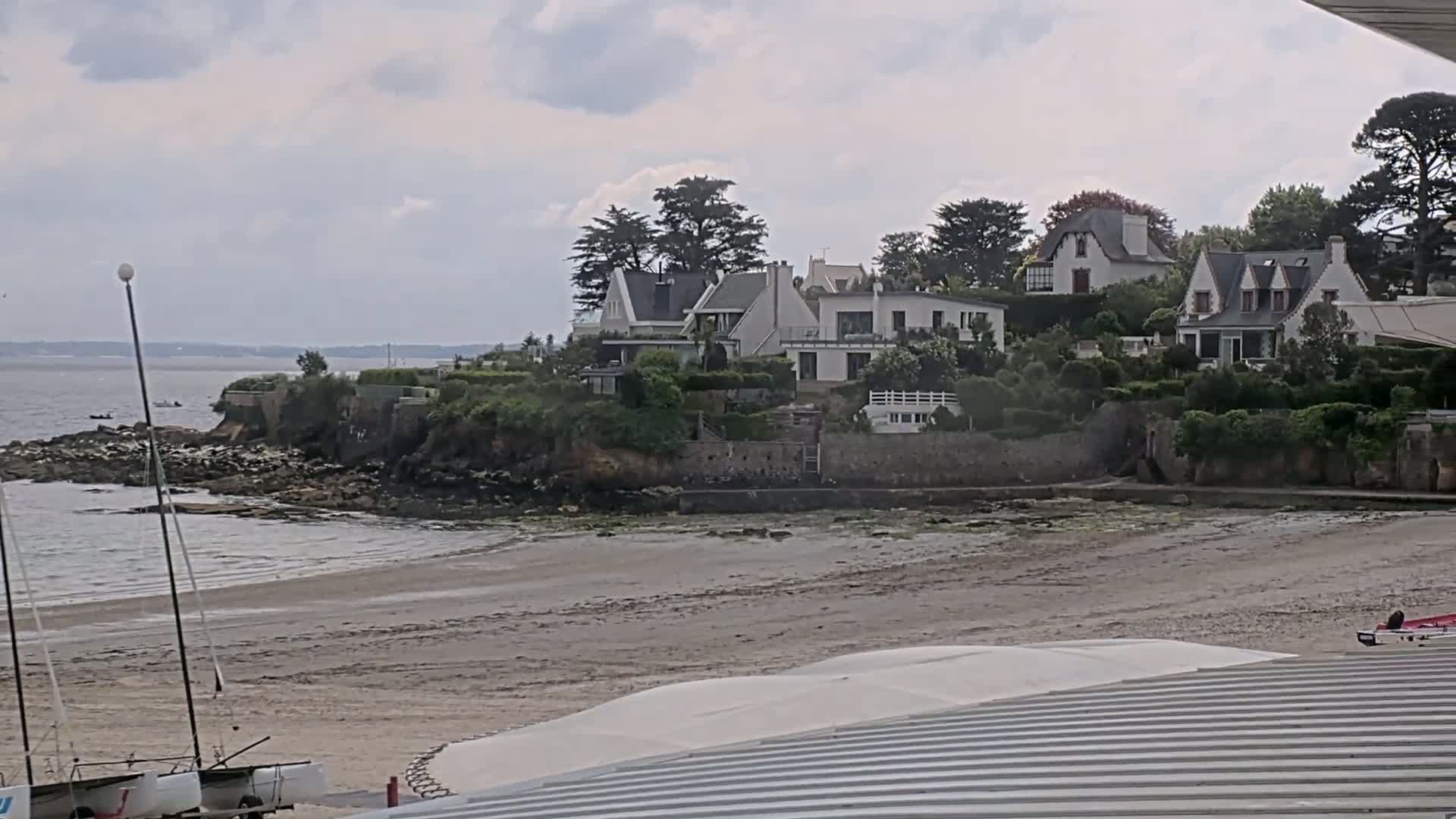 A sandy beach with several sailboats and a cluster of white houses nestled on a rocky hillside overlooking calm water under a cloudy sky.