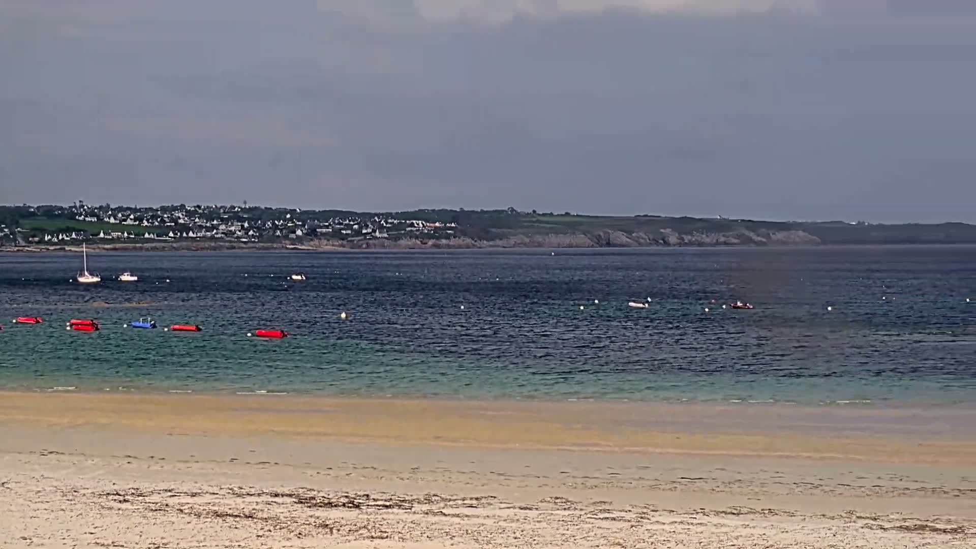 A sandy beach slopes gently to a calm, teal-colored sea, dotted with small boats and buoys, under a cloudy sky, with a distant town visible on the horizon.