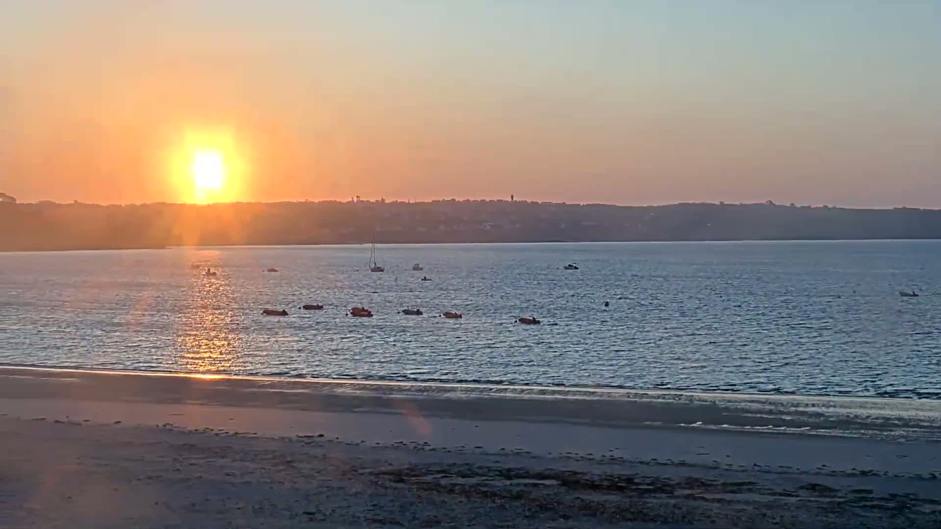 A calm body of water with several small boats sits before a sandy beach at sunset, with a distant shoreline visible in the background.