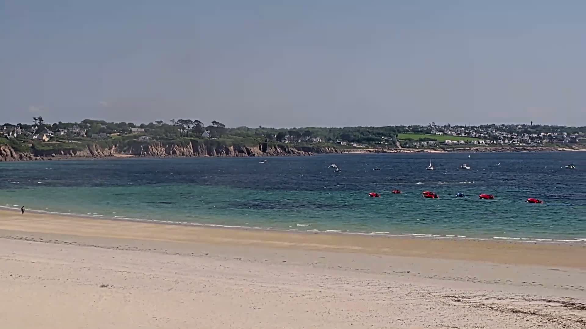 A sandy beach stretches toward a calm, teal-colored bay under a clear blue sky, with small boats scattered on the water and a distant town visible across the bay.