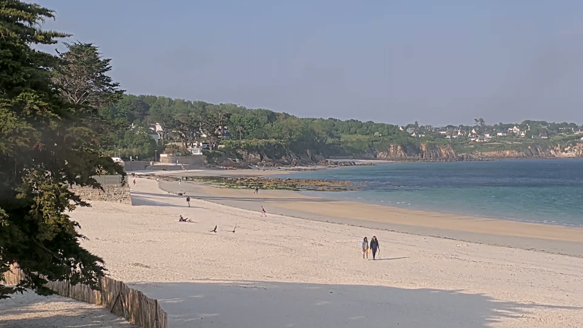 A sandy beach stretches along a calm, light-blue ocean under a clear sky, with a few people walking and relaxing on the sand and green hills and houses in the background.