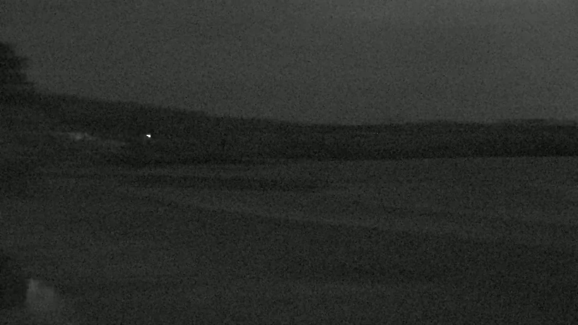 A dark, grainy nighttime image shows a flat, dark landscape with a single faint light in the distance.