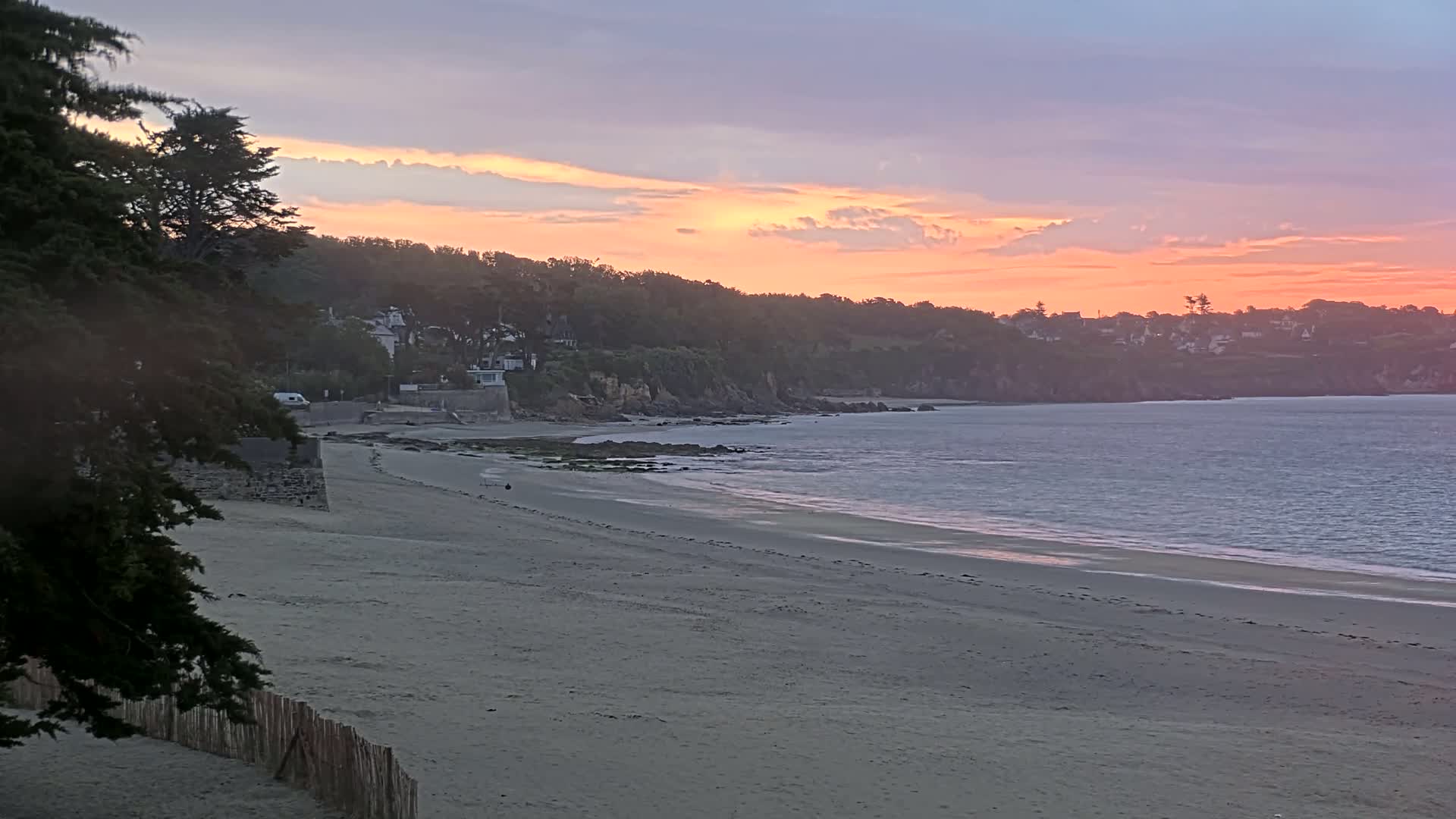 A sandy beach slopes gently toward calm ocean water under a sunset sky with soft pink and orange hues.