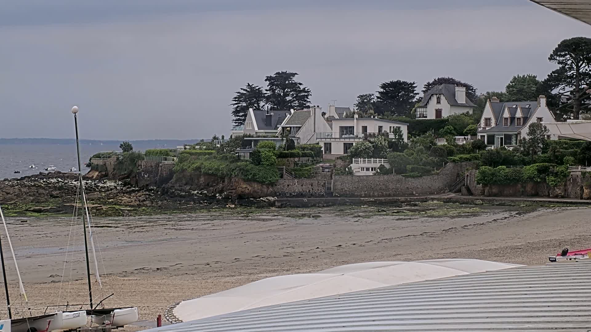 A sandy beach with two sailboats is visible in the foreground, while a cluster of houses sits atop a rocky outcropping overlooking a calm sea under an overcast sky.