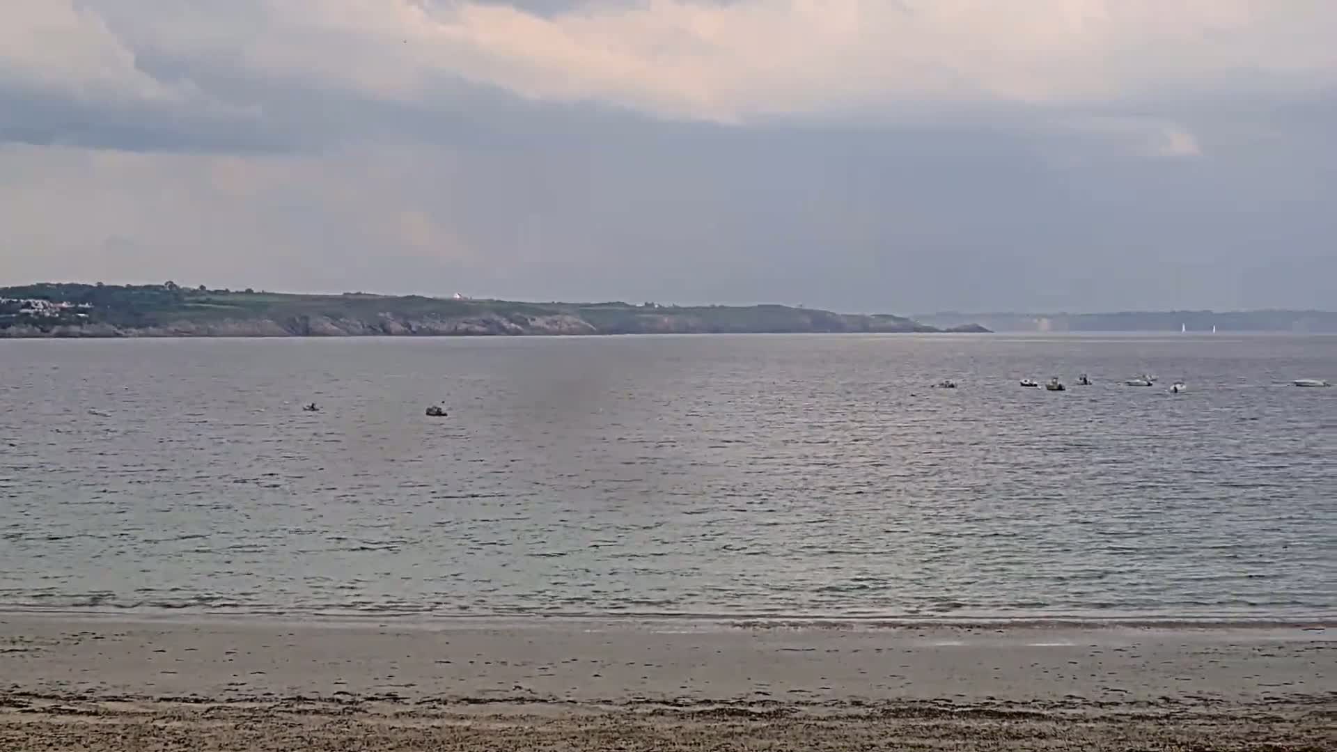 A sandy beach stretches toward a calm sea dotted with small boats under a cloudy sky.