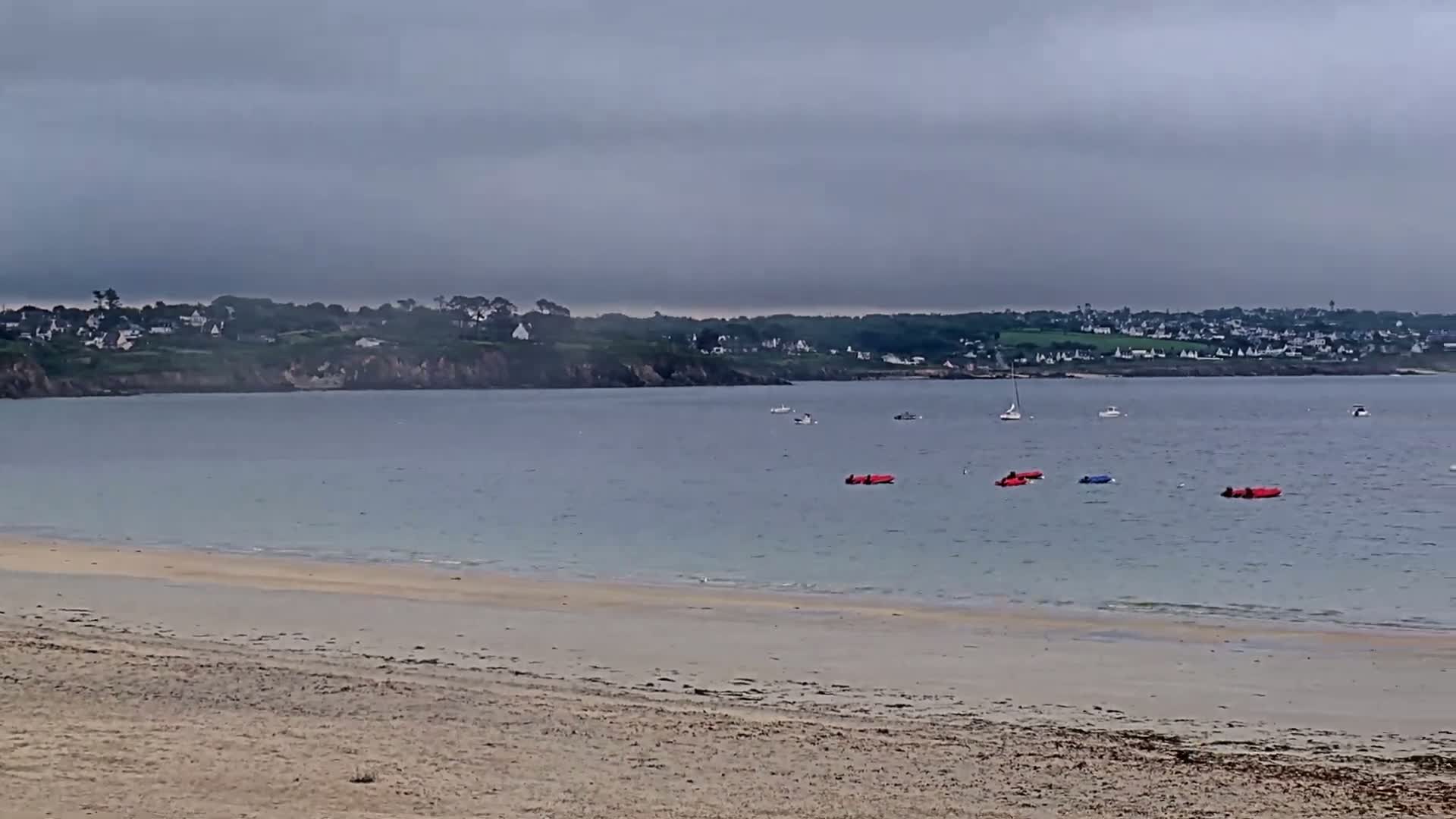 A sandy beach stretches to a calm bay under an overcast sky, with several small boats and inflatable kayaks visible on the water and a distant shoreline with houses.