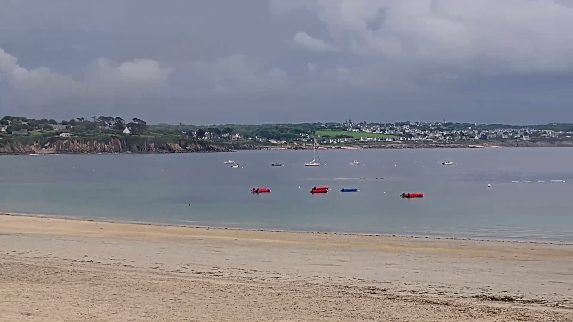 A sandy beach stretches to a calm bay under a cloudy sky, with several small boats and inflatable rafts in the water and a distant shoreline with buildings visible.