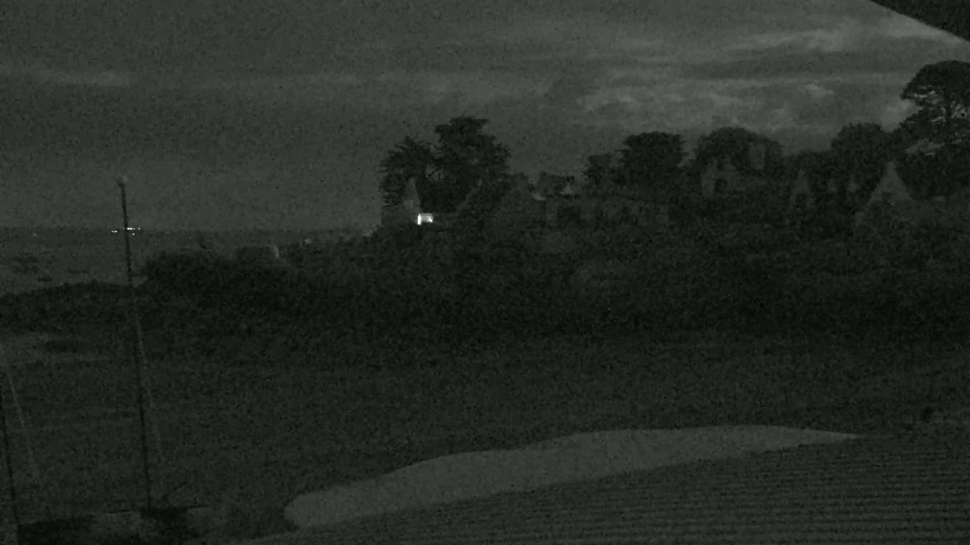 A nighttime view shows a dark, tree-lined landscape with a few lit windows in the distance, under a cloudy sky.