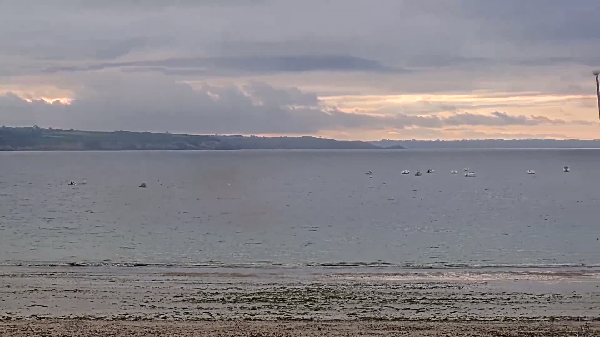 A calm, overcast day with a subtle sunset shows a sandy beach, a body of water with several small boats and birds, and a distant shoreline.